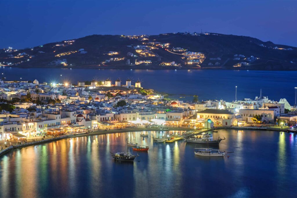 destination with famous windmills, and port with boats and yachts illuminated in the evening blue hour . Mykonos, Cyclades islands, Greece