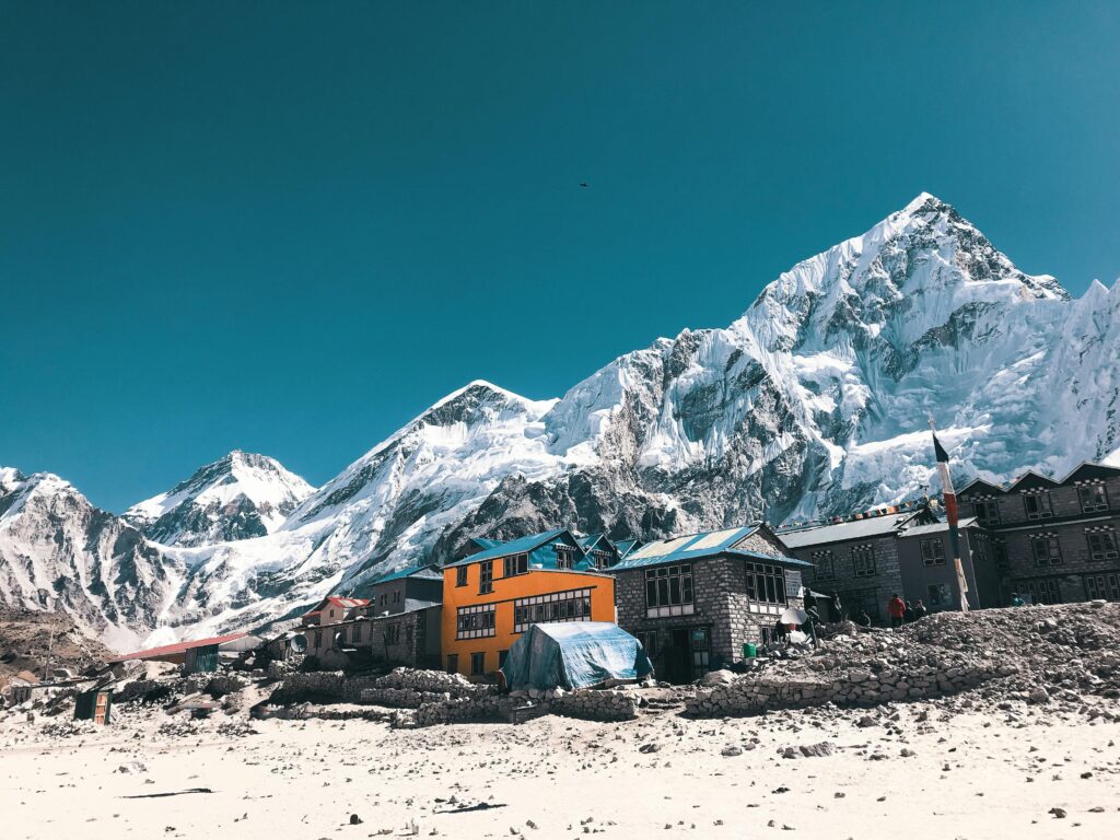 Village at the base of snow-covered mountains with stone buildings and a bright orange building under a clear blue sky, highlighting the journey to Climb Mount Everest.